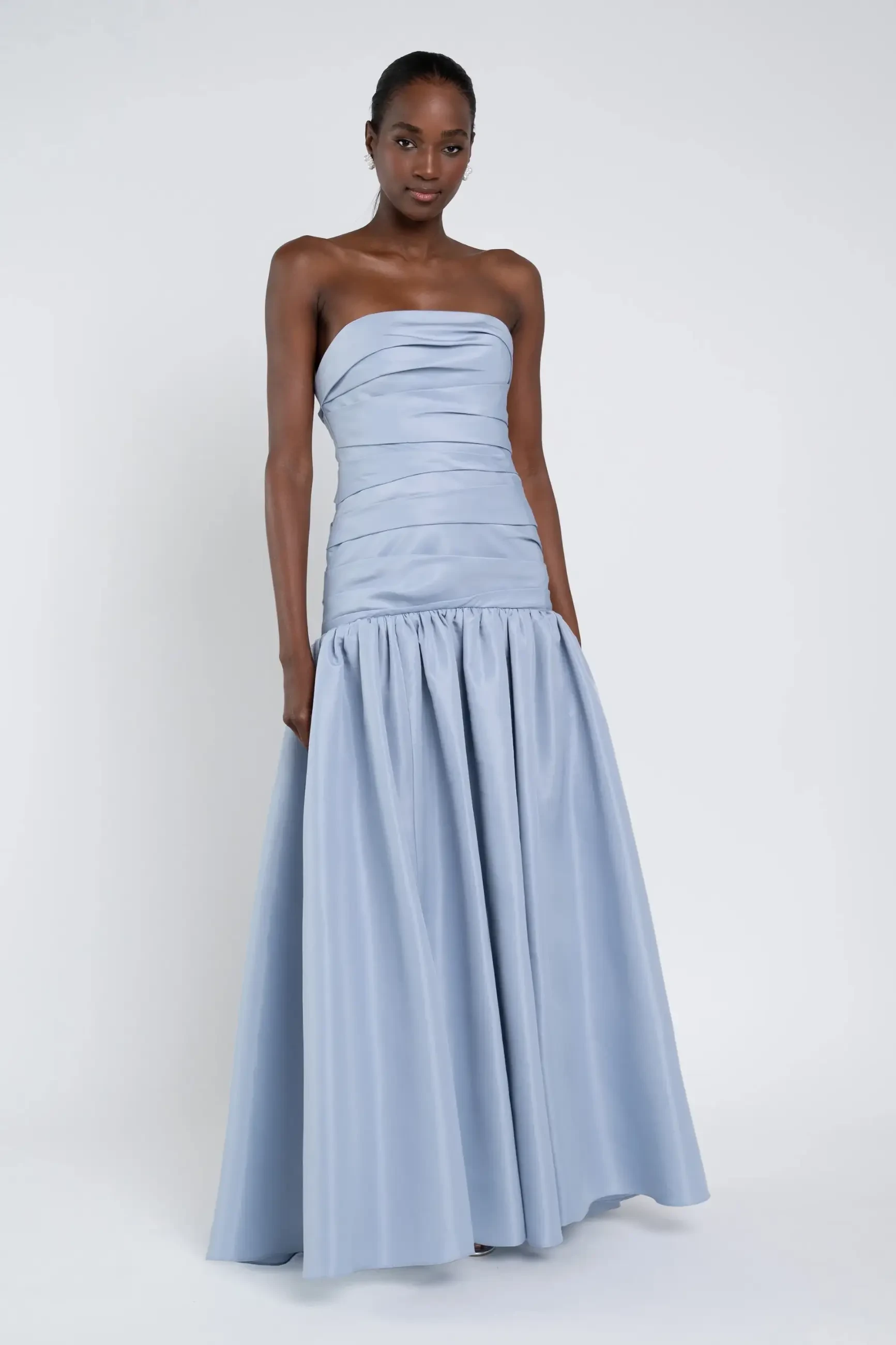 A woman in an elegant light blue strapless gown with a fitted bodice and flowing skirt stands against a white background, conveying grace and sophistication.