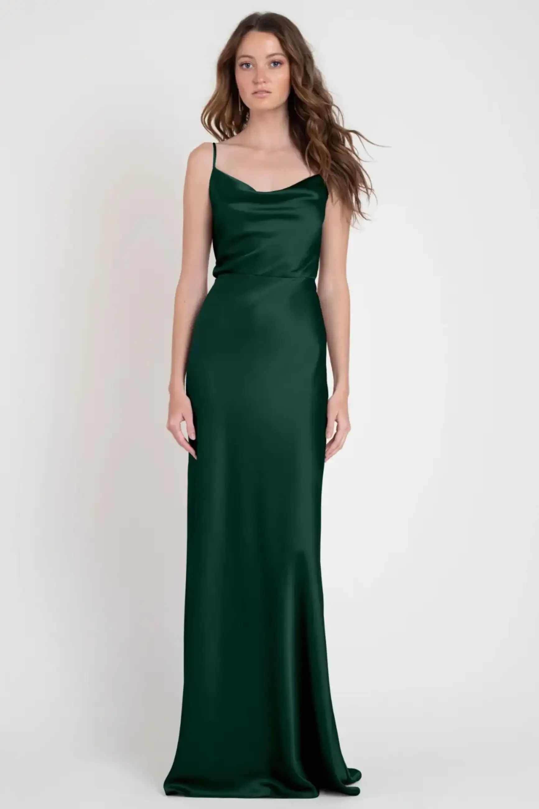 Woman wearing an elegant, floor-length, dark green satin gown with thin straps, standing against a plain white background, projecting a sophisticated and serene aura.