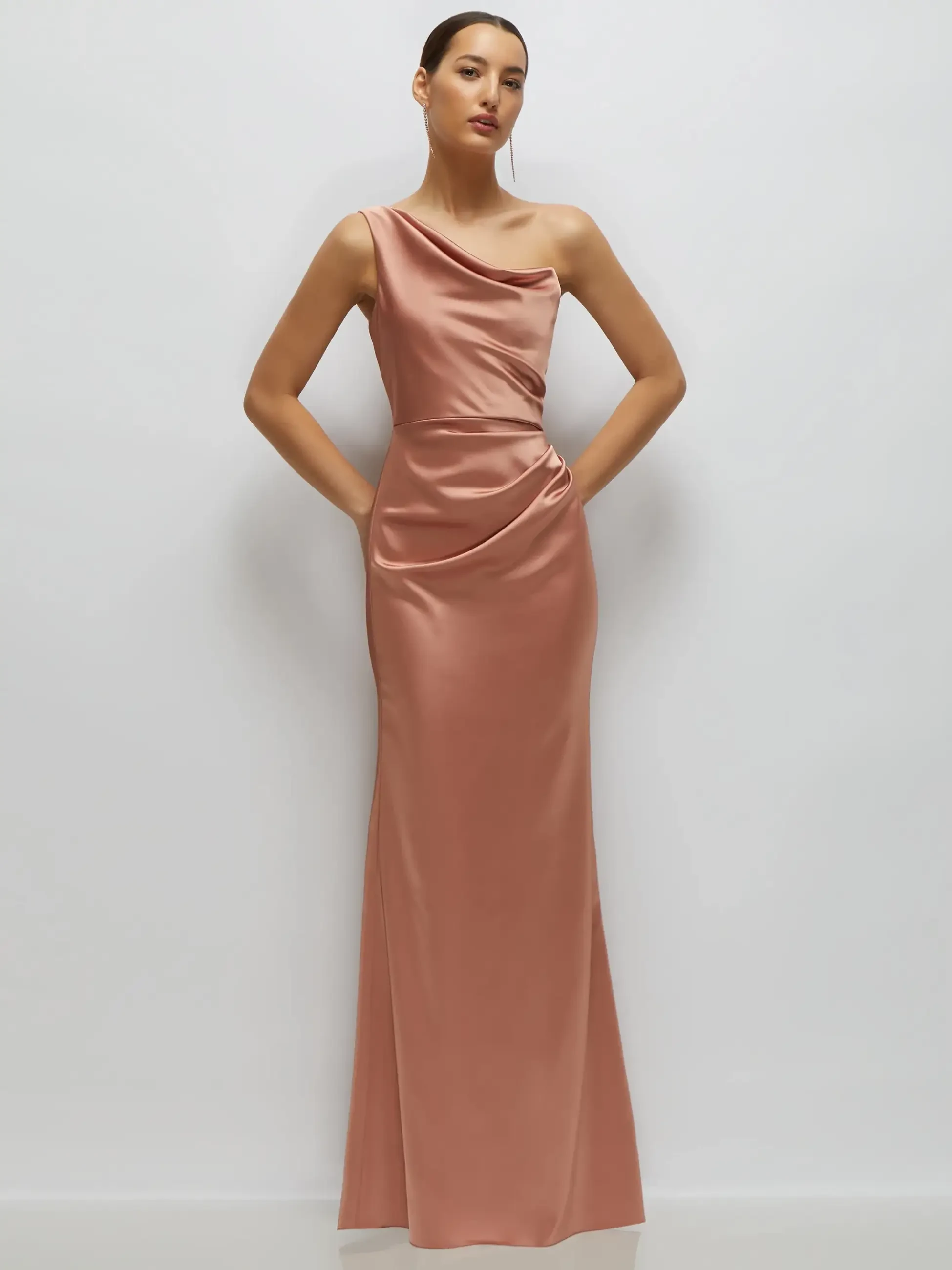A model wearing a one-shoulder, floor-length satin gown in a soft pink color, displaying a draped design with pockets.