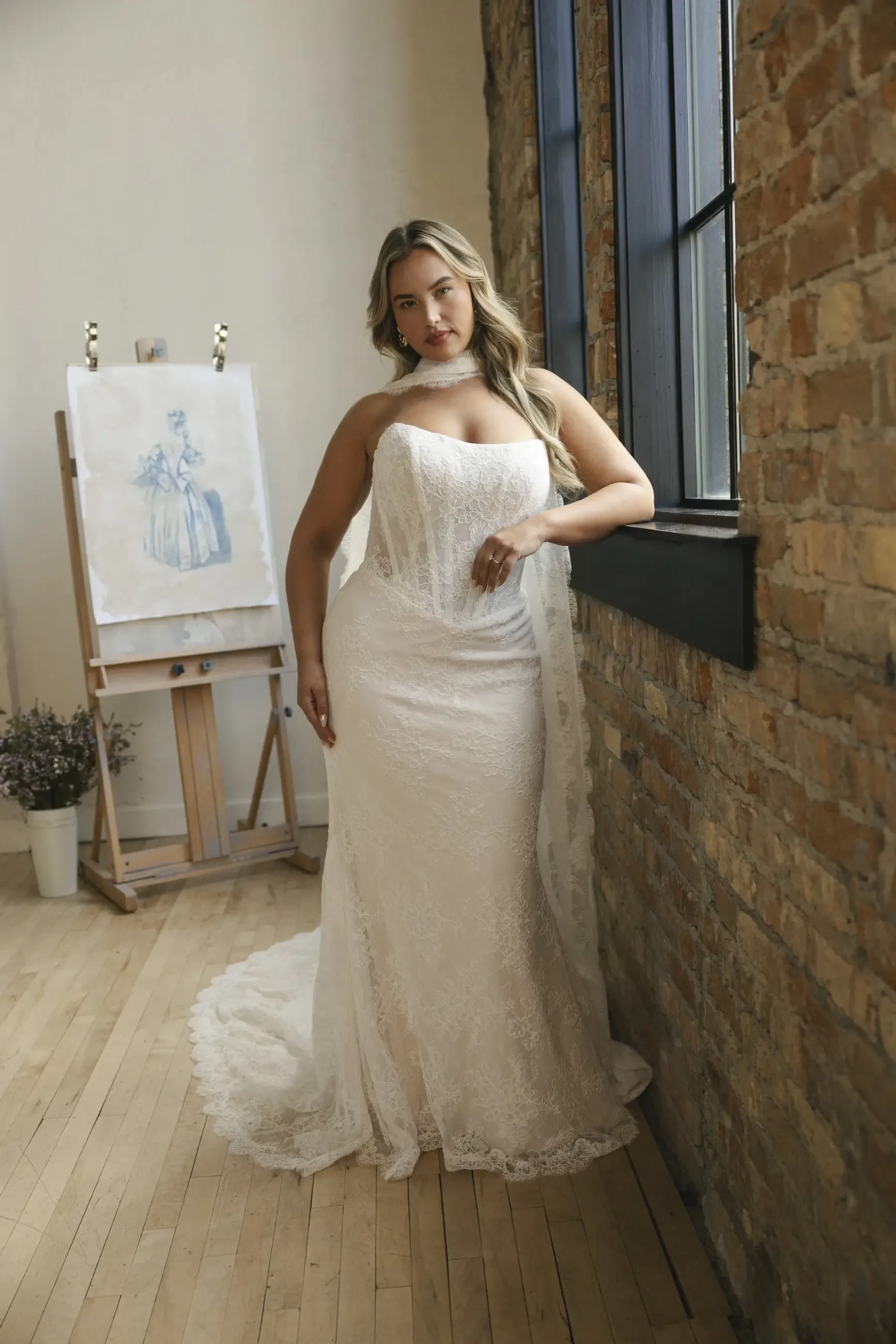 A woman in a lace wedding dress leans against a rustic brick wall, exuding elegance. An easel with a gown sketch and a potted plant are in the background.