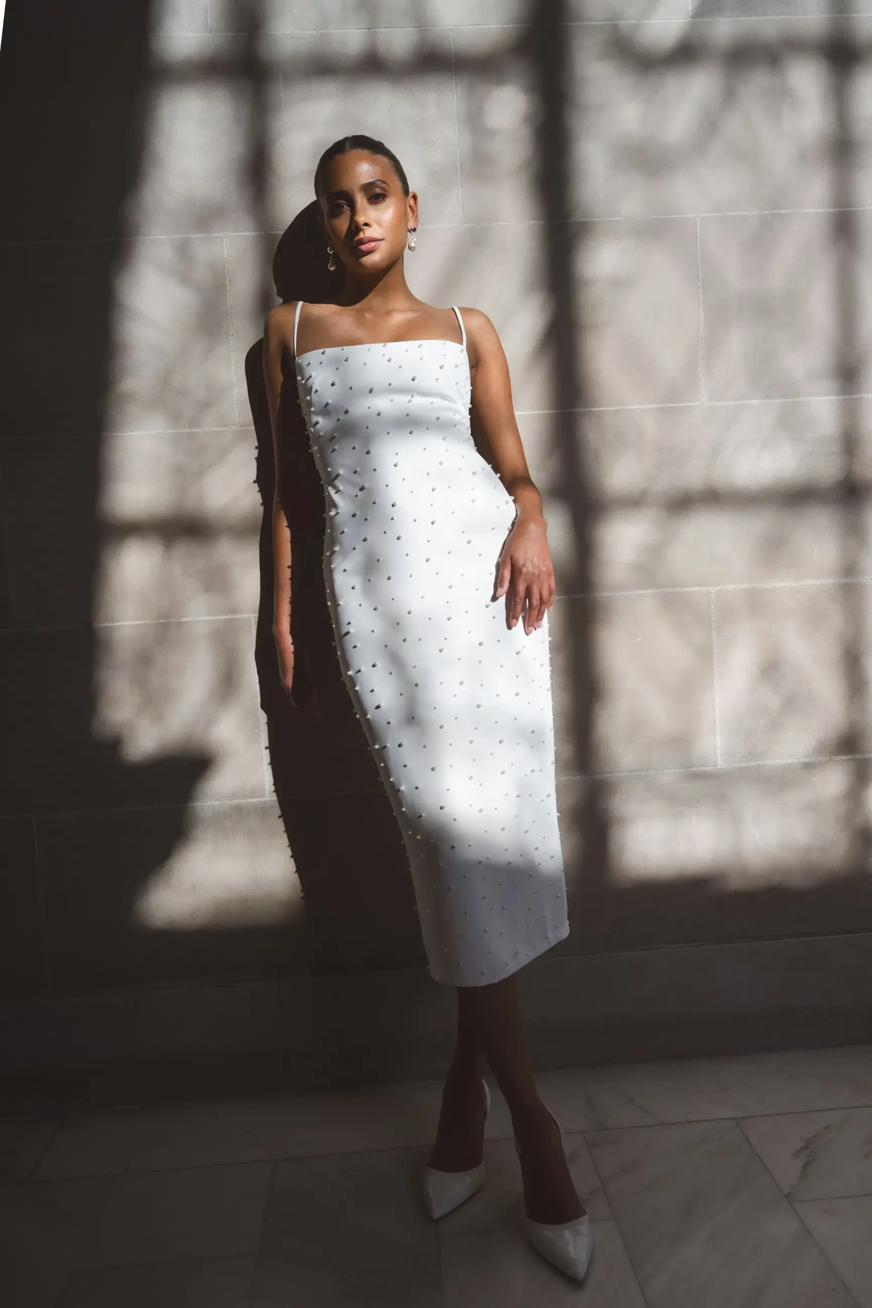 Model is wearing a sheath midi wedding dress with a square neckline and spaghetti straps with pearls encrusted all over the dress
