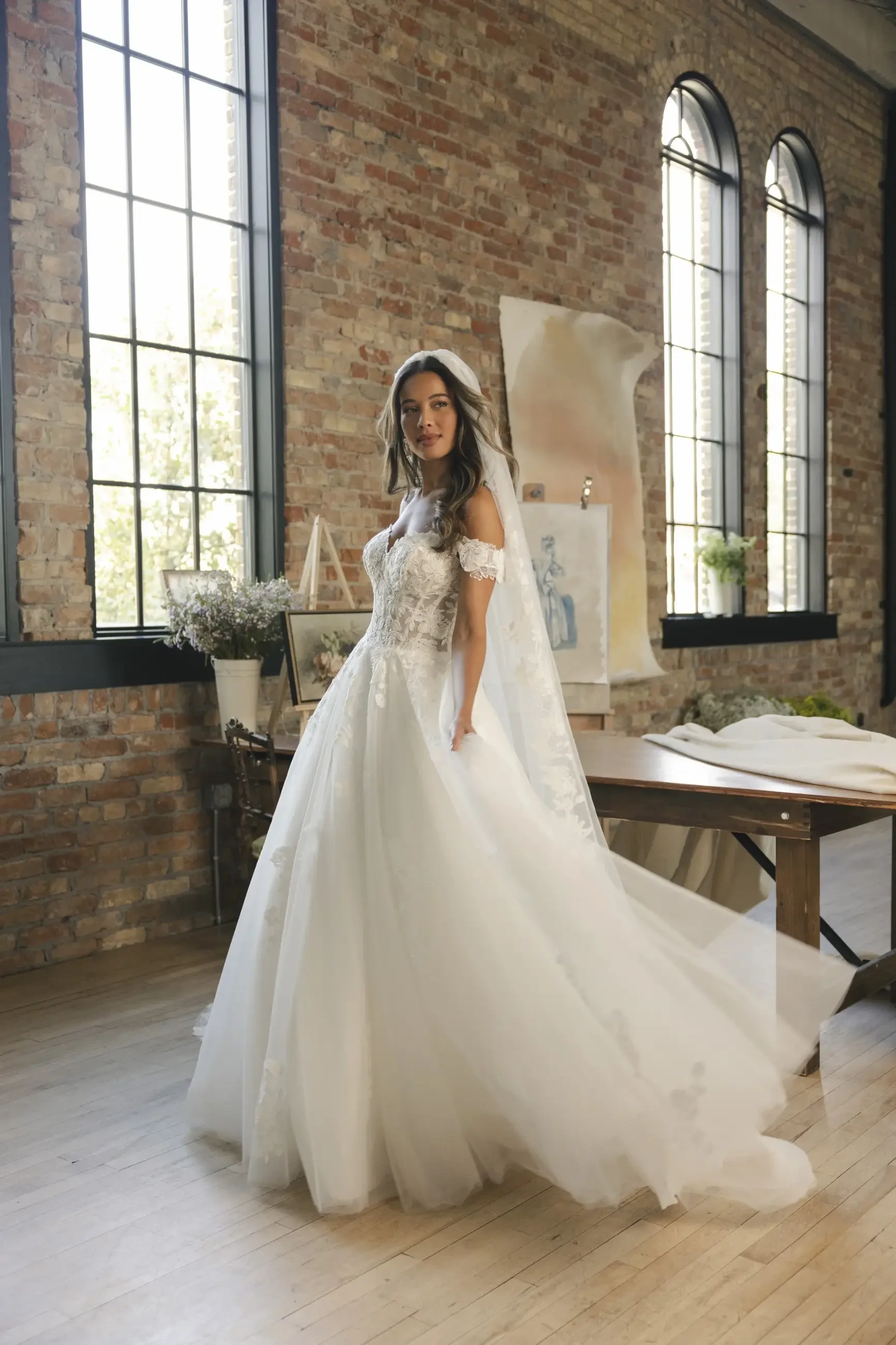 Bride in an elegant white gown with lace details and a flowing veil stands in a sunlit room with large brick walls and tall windows, exuding romance.