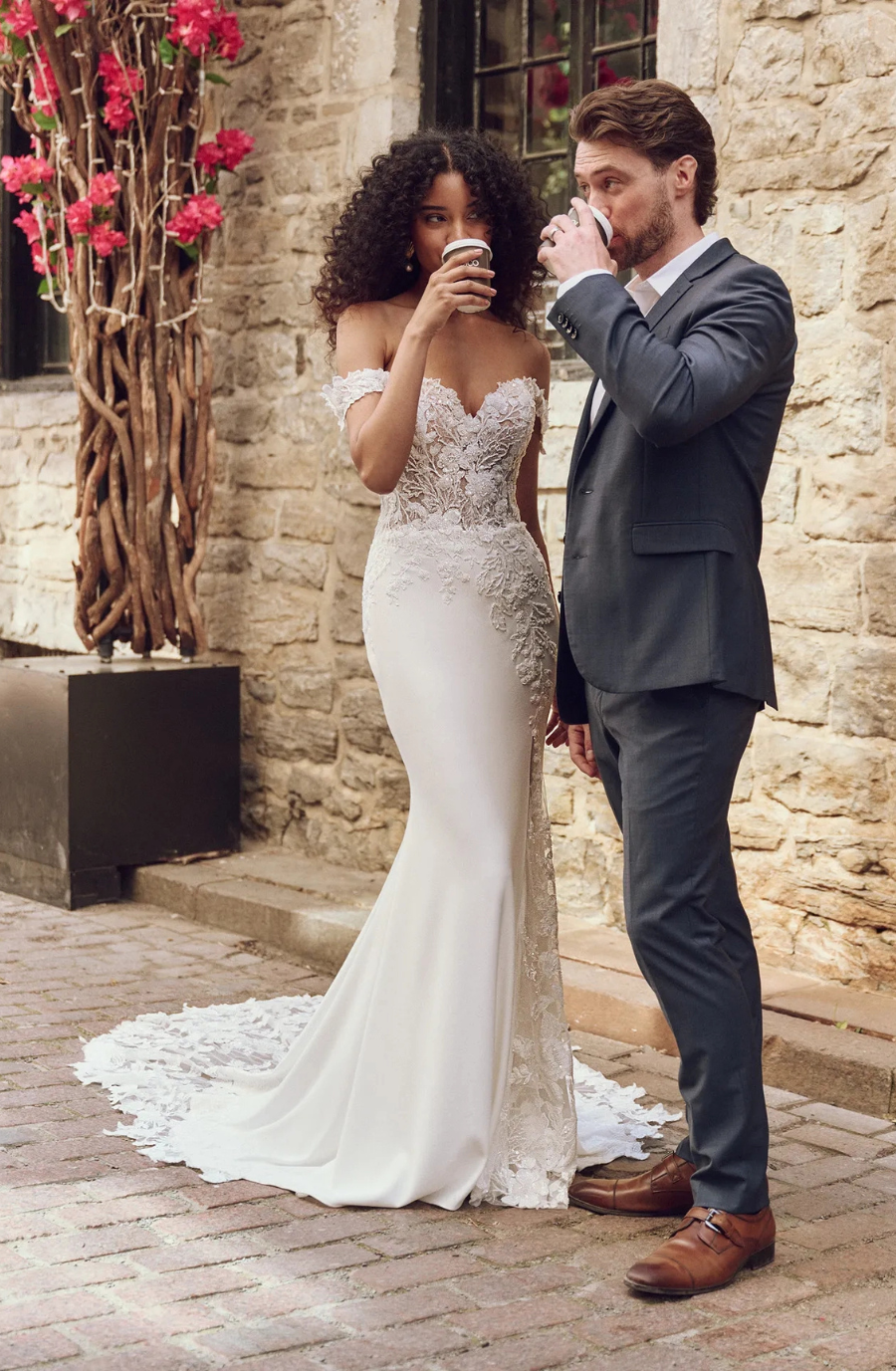 Bride in an elegant lace wedding gown and a groom in a navy suit sipping coffee outdoors, standing by a stone wall adorned with pink flowers.