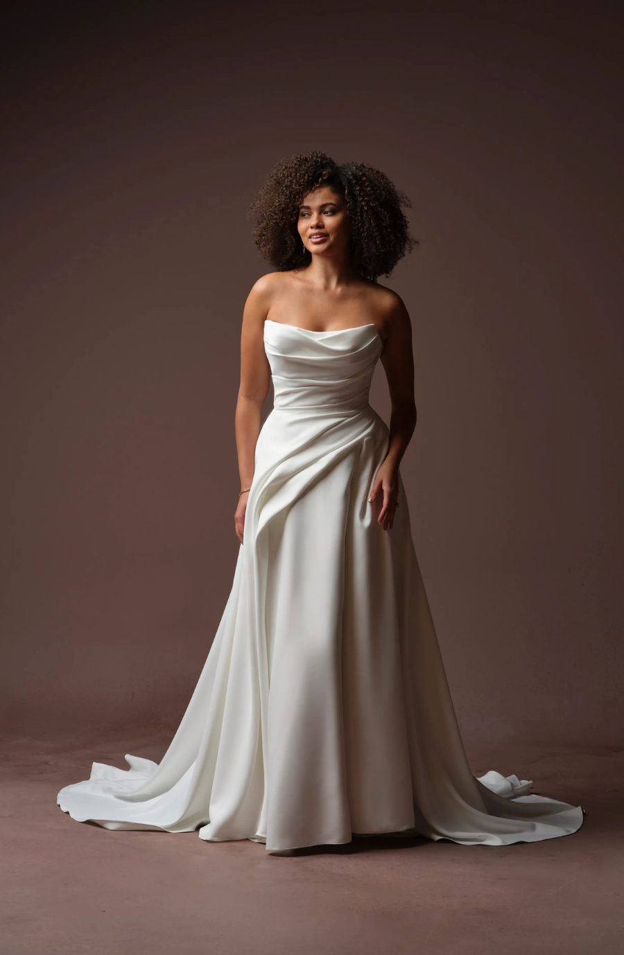 A woman in a strapless white gown stands confidently against a neutral background, with her curly hair styled naturally.