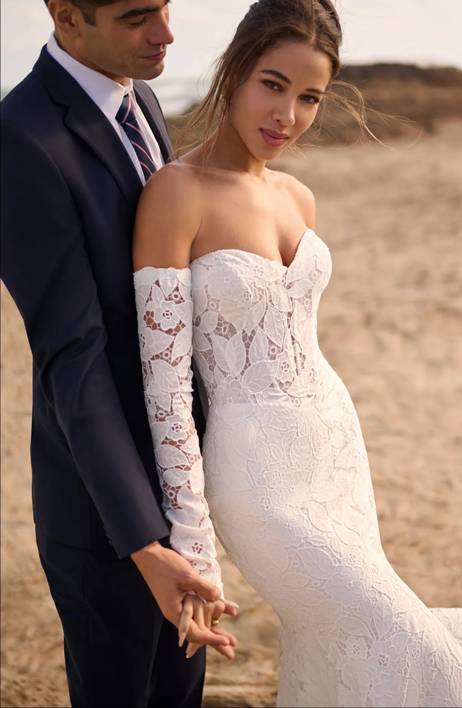 A couple stands on a beach, holding hands. The woman wears an elegant, off-shoulder lace wedding dress, while the man is in a navy suit. They exude joy.