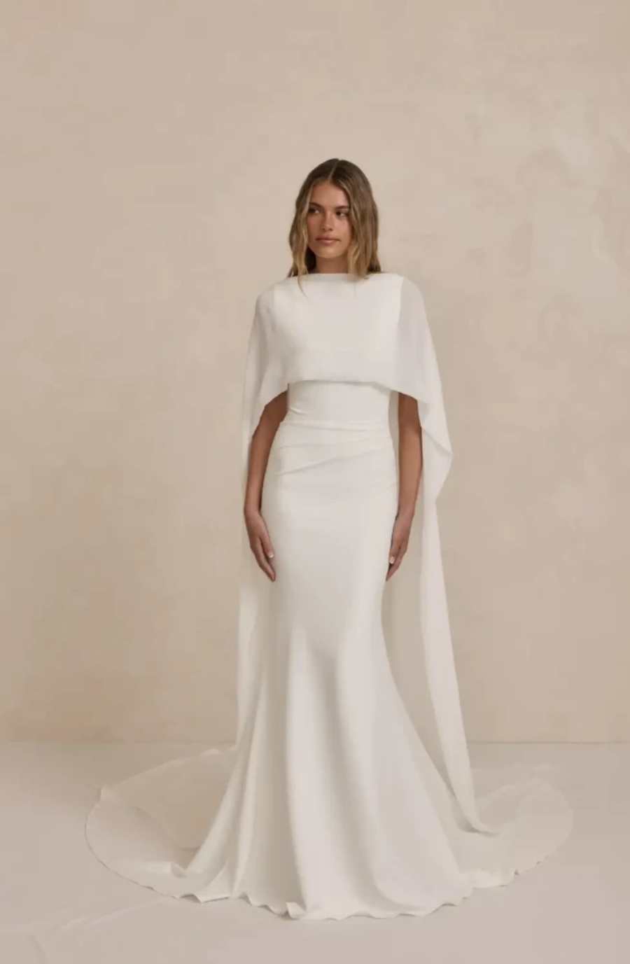 A woman stands in an elegant, fitted white gown with a flowing cape. The minimalist backdrop emphasizes the dress's sleek, sophisticated design.