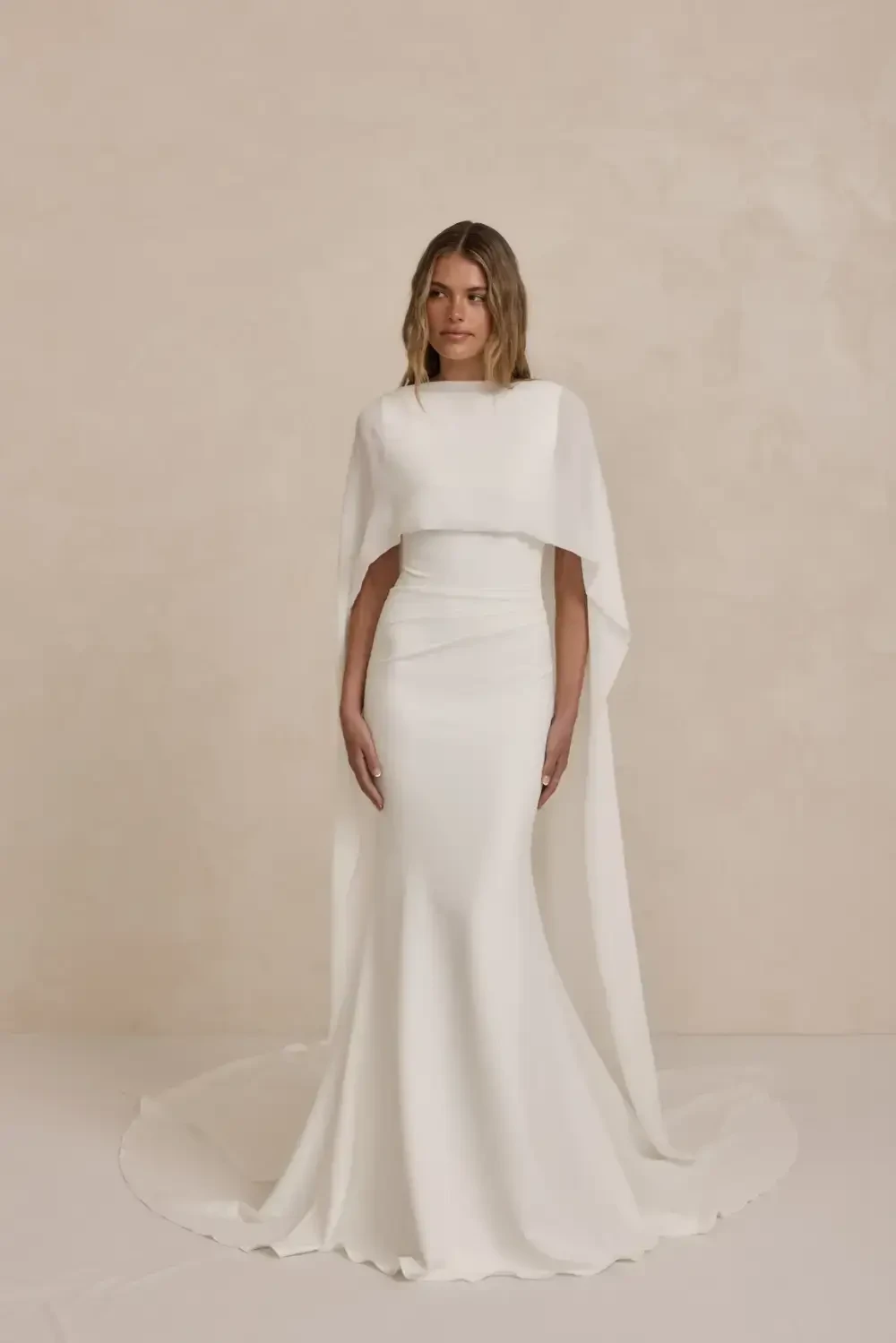A woman wearing a white gown with a cape, standing against a neutral background.