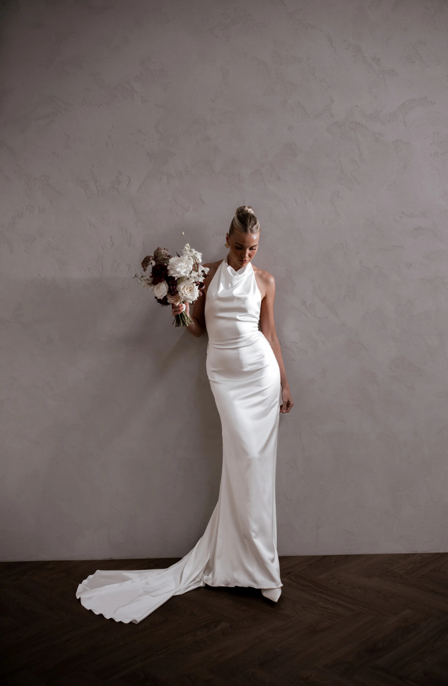 Bride in a sleek, white satin gown with a halter neck holds a bouquet of white and dark flowers, standing against a textured gray wall. Elegant and serene.