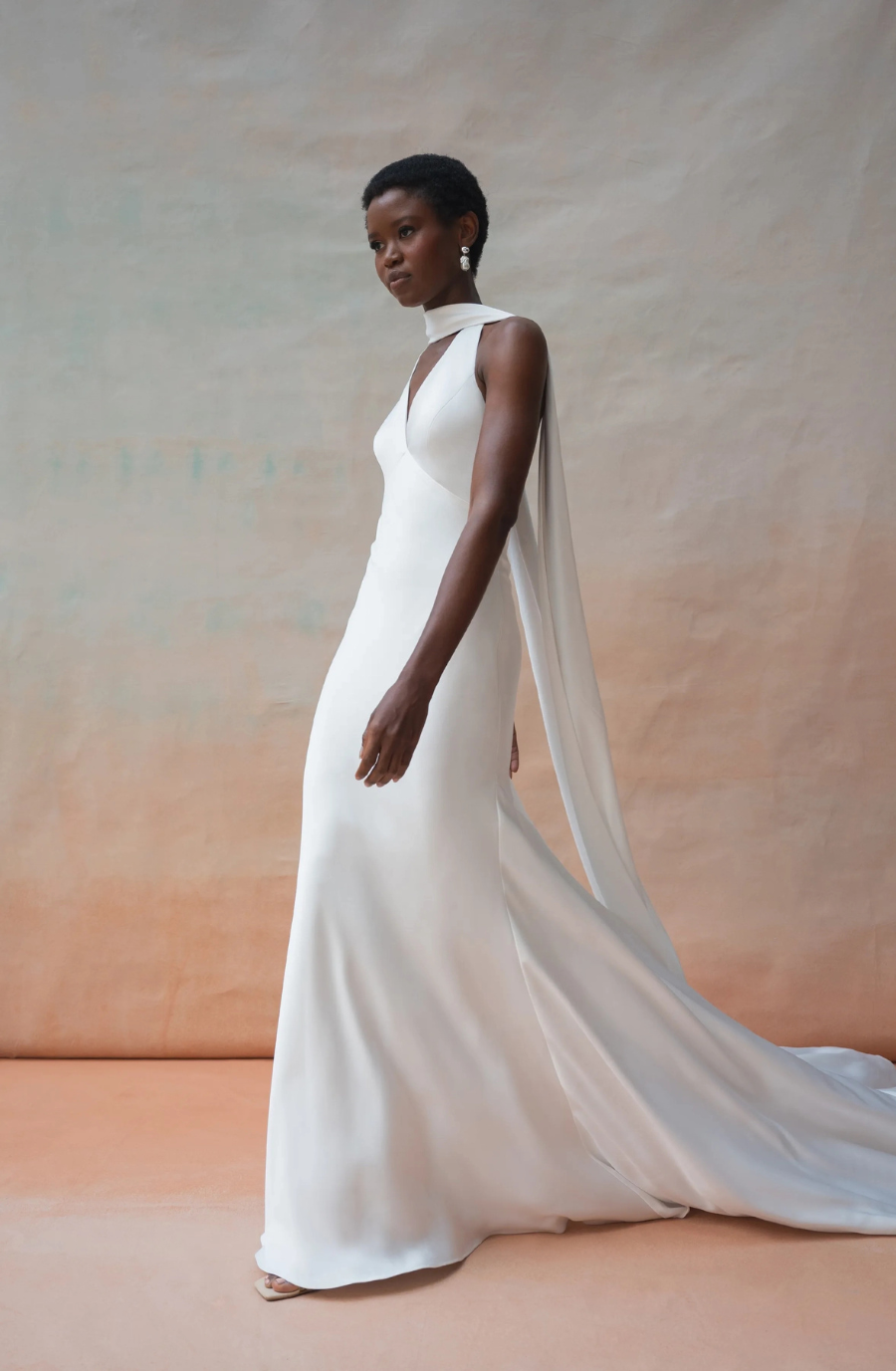 A woman in a flowing white gown with a high neckline and cape stands elegantly against a beige background. Her expression is poised and serene.
