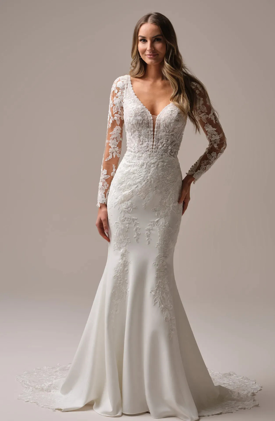 A woman wears a white wedding dress with lace sleeves and a deep V-neck. The dress features intricate floral patterns and a flowing train, conveying elegance.