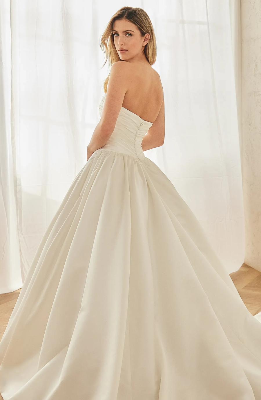 Woman in an elegant strapless white wedding gown, standing in a softly lit room with sheer curtains. She gazes over her shoulder, exuding grace.