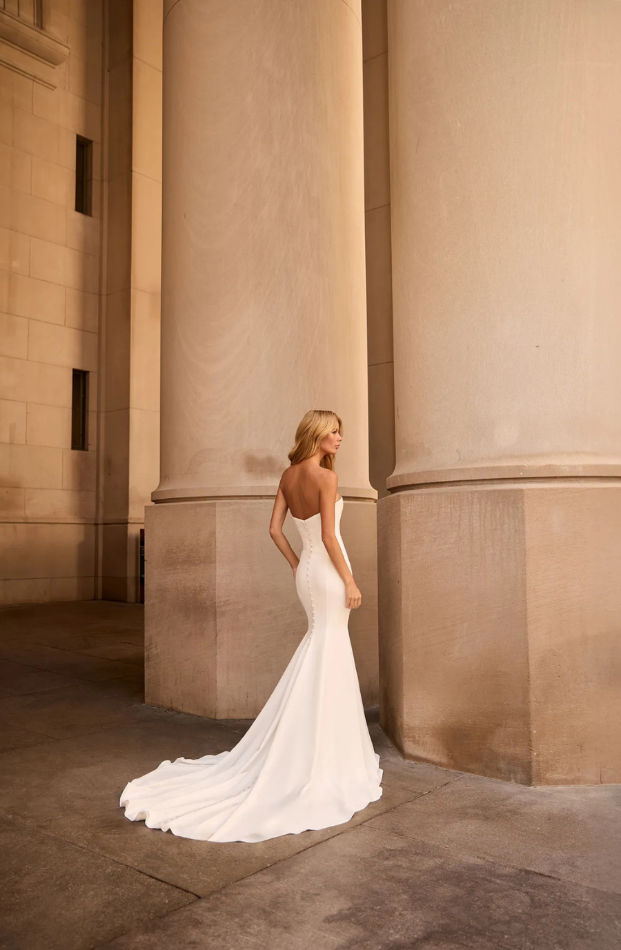 A woman wearing a sleek, strapless white wedding dress stands elegantly near large stone columns.
