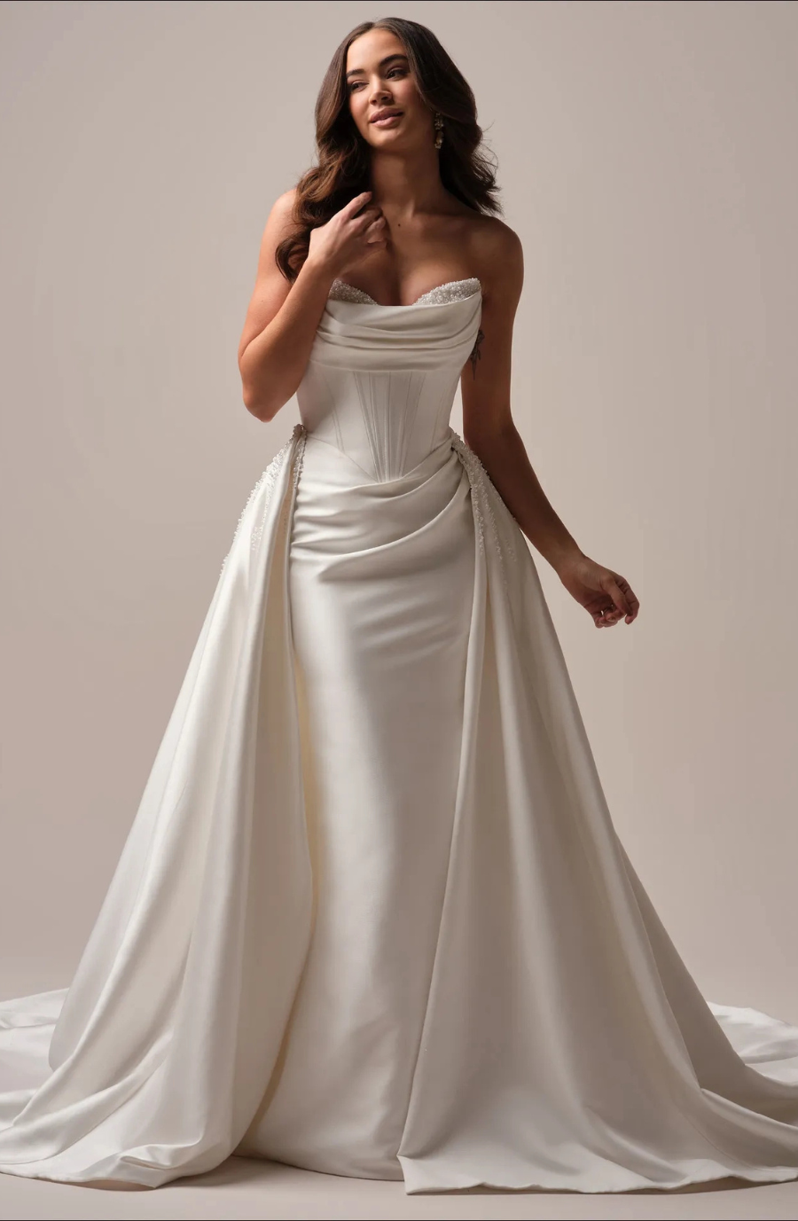Elegant woman in a strapless white satin wedding gown with a draped bodice and long train, exuding grace and confidence against a neutral background.