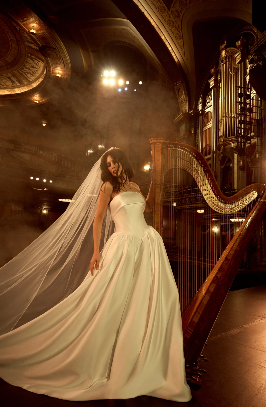 A bride in a white gown with a long veil stands near a harp in a grand venue, with dramatic lighting and a misty atmosphere.