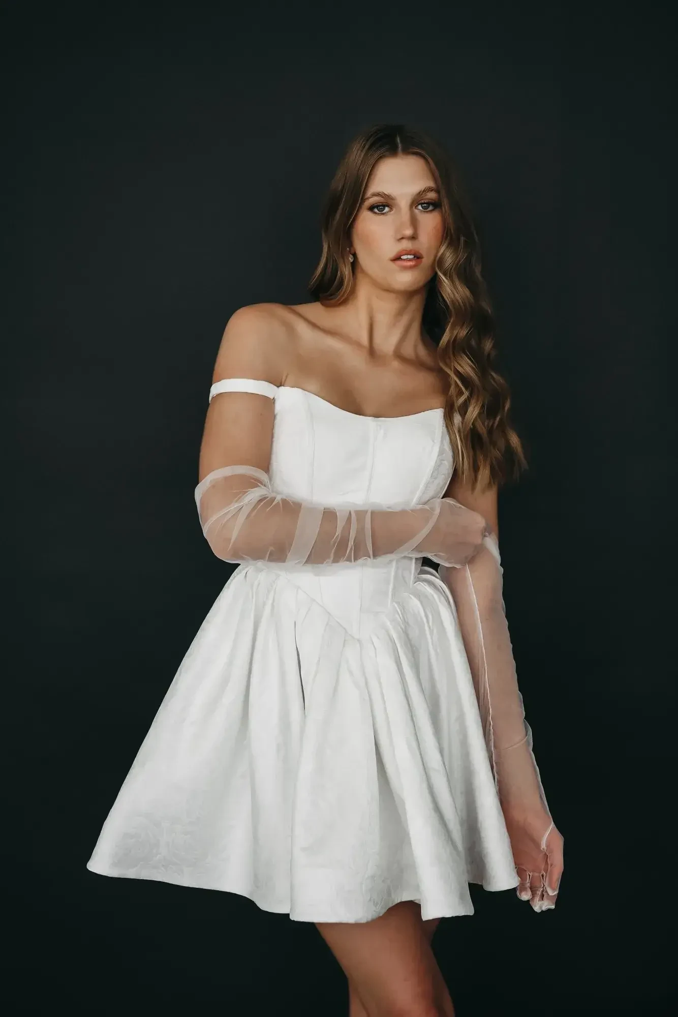 A woman in a white off-shoulder dress with sheer gloves stands against a dark backdrop. Her expression is poised, and her long, wavy hair cascades down.