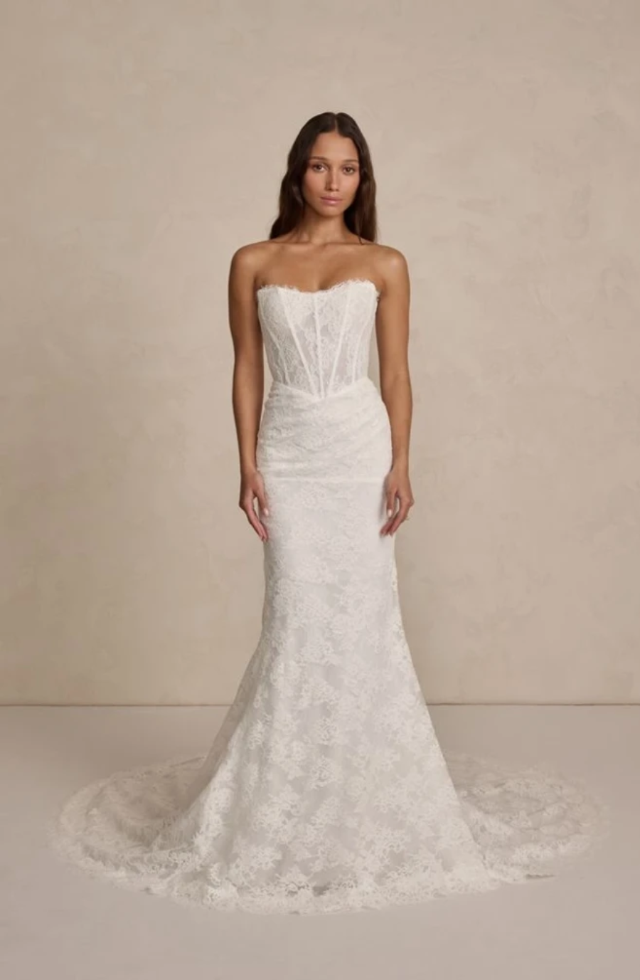 A woman in a strapless, lace wedding gown with a fitted bodice and long train stands against a soft beige background, conveying elegance and simplicity.