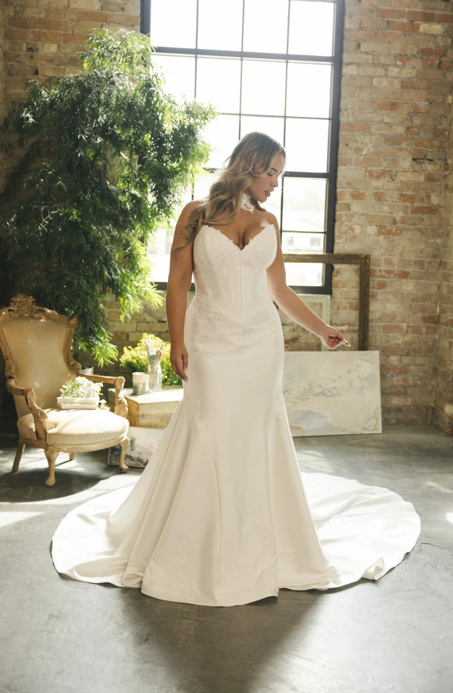 Bride in elegant white gown with lace details stands in sunlit room. Brick walls, large window, vintage chair, and greenery create a romantic ambiance.