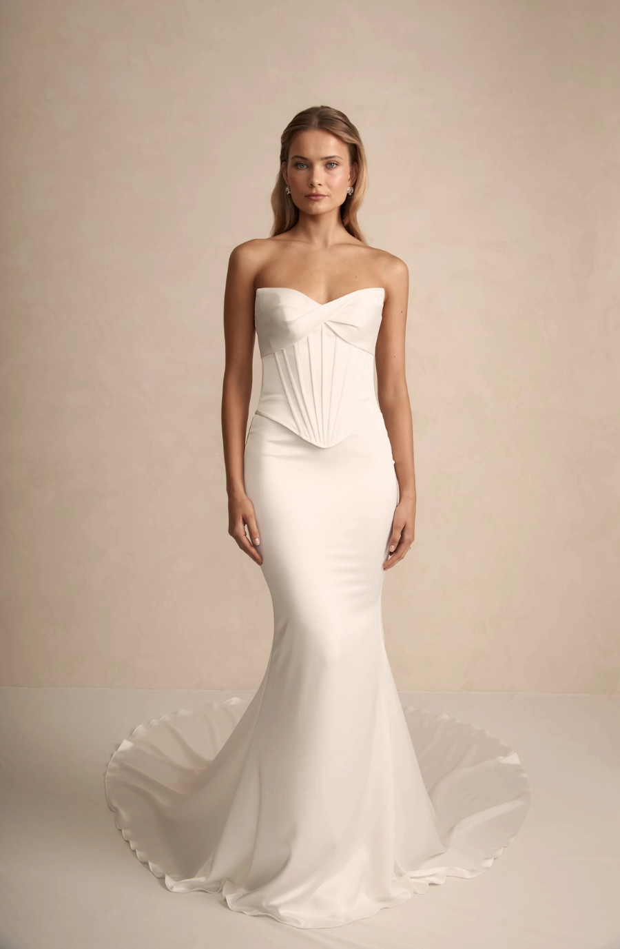 A woman stands wearing a strapless, fitted white wedding gown. The dress has a structured bodice and a mermaid silhouette, creating an elegant and serene look.